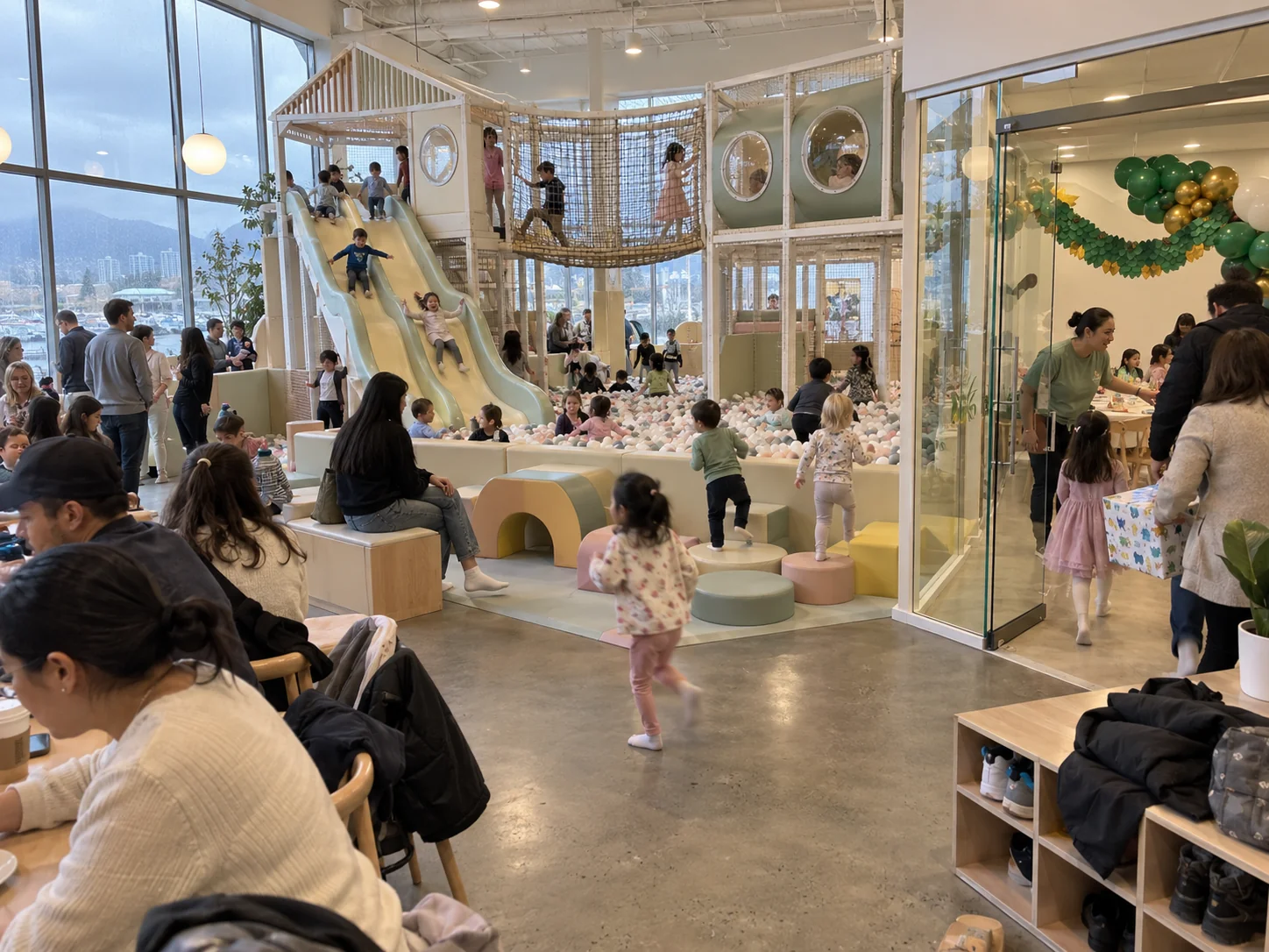 A busy indoor playground with slides, nets and a ball pit, and a glass-walled party room with green and gold balloons where a host welcomes a family